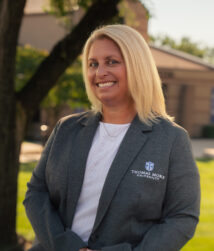 Director of Admissions Trinity Walsh on a sunny day sitting on a bench with the Saints Center behind her.