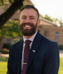 A photo of Josh Ostertag, the Director of the Bishop Foys Scholars Program and Assistant Campus Minister. He is sitting on a bench in front of the chapel, and the Saints Center is in the background.