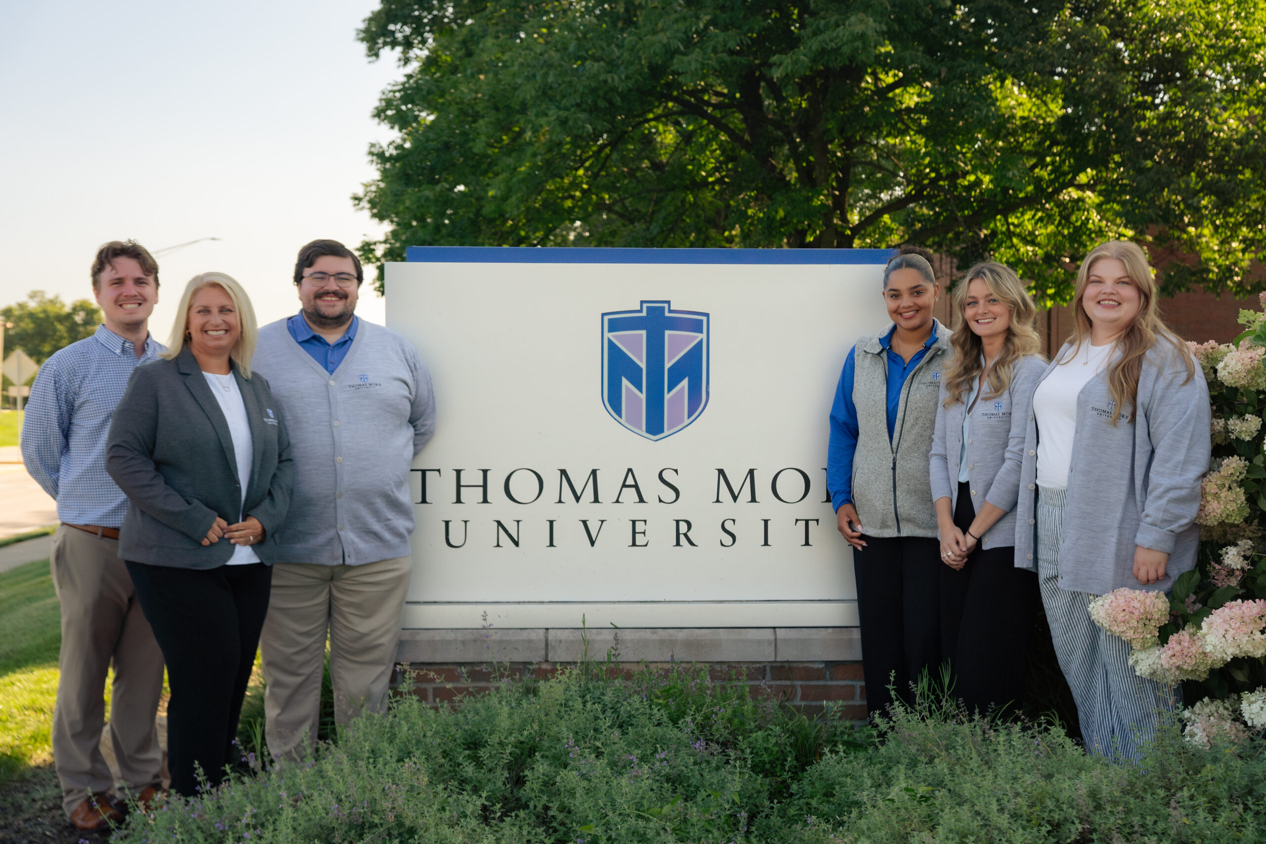 The Thomas More University Admissions team standing in front of a sign on campus with the University's name. They are surrounded by vibrant plants and large trees and blue skies are behind them.