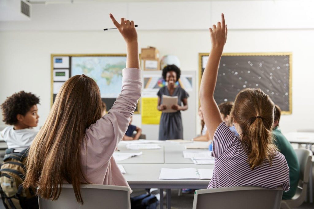 students raising hands in a classroom with a smiling teacher in the background