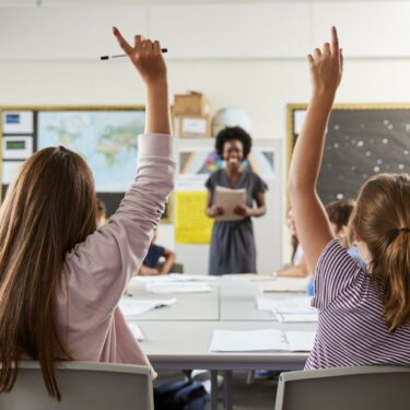 students raising hands in a classroom with a smiling teacher in the background